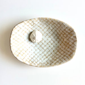 May include: A white ceramic bowl with a textured, diamond-shaped pattern. The bowl is oval-shaped and has a slightly raised rim.