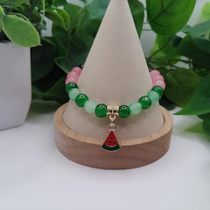 May include: A bracelet with pink and green beads, and a watermelon charm. The charm is red and green, and the bracelet is displayed on a wooden stand. The beads are approximately 0.8 cm in diameter.