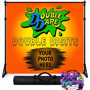 May include: Orange and yellow backdrop with the text "DOUBLE DARE" and "DOUBLE DIGITS". The backdrop has a space for a photo and is supported by a black stand. A black carrying case is at the bottom.
