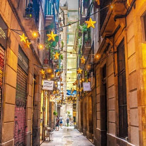 Puede incluir: Un callejón estrecho y empedrado en Barcelona, España, decorado con luces de cadena y estrellas doradas. El callejón está bordeado de edificios, algunos con tiendas y restaurantes. Los edificios están pintados en tonos cálidos y terrosos.