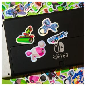 May include: A collection of colorful, holographic stickers featuring various weapon designs, including a blaster, a roller, and a paint gun. The stickers are scattered on a black Nintendo Switch console, with the Nintendo Switch logo visible.