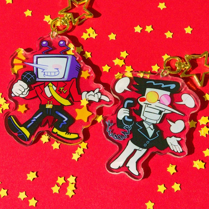 May include: Two acrylic keychains featuring cartoon characters on a red background scattered with yellow star confetti. One keychain depicts a character with a TV head and a microphone, the other a character with a long nose and a phone.