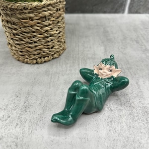 May include: A green ceramic figurine of a reclining elf with a mischievous expression. The elf is wearing a green hat and green clothing.
