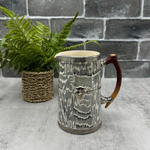 Peut inclure: Pichet décoratif avec un bord et une poignée argentés, présentant un motif gris et blanc. La poignée est marron et dorée. Un petit fermoir argenté avec un visage est sur le devant. Une petite plante en pot est en arrière-plan.