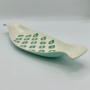 May include: A white ceramic serving dish with a green and yellow geometric pattern. The dish has a curved shape and a handle on one end.