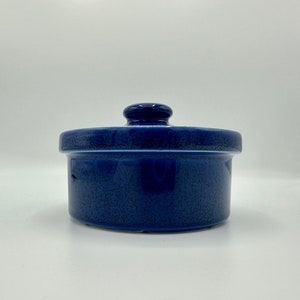 May include: A small, round, blue ceramic container with a lid. The container has a speckled texture and a smooth, rounded lid.