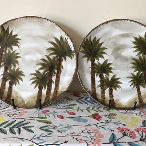 May include: Two ceramic plates with a tropical palm tree design. The plates have a white background with brown and green palm trees. The rims are brown. The plates are displayed on a floral patterned tablecloth.