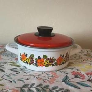 May include: A white enamel pot with a red lid and black handle. The pot features a floral design in orange, yellow, and green. The pot has two white handles and is sitting on a floral patterned tablecloth.
