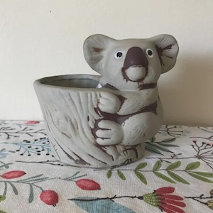 May include: A gray ceramic koala planter. The koala has a brown nose and is sitting inside a tree trunk-shaped pot. The planter is on a floral patterned surface.