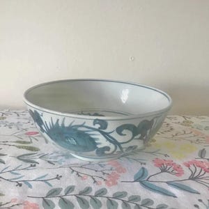 May include: A white ceramic bowl with a blue floral design. The bowl has a wide rim and a rounded shape. The floral pattern is painted in shades of blue and covers the exterior of the bowl.