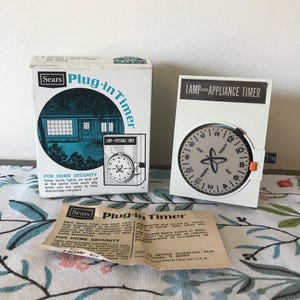 May include: A vintage Sears Plug-in Timer set, including a box and timer unit. The box features a blue and white illustration of a house. The timer has a white face with a black dial and orange accents. The text "LAMP AND APPLIANCE TIMER" is visible.