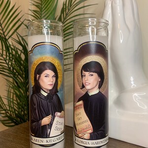 May include: Two white prayer candles with images of women and text. The candle on the left has an image of a woman with dark hair and the text "Karen Kilgariff Stay Sexy". The candle on the right has an image of a woman with dark hair and the text "Georgia Hardstark Don't Get Murdered Eat Pizza".