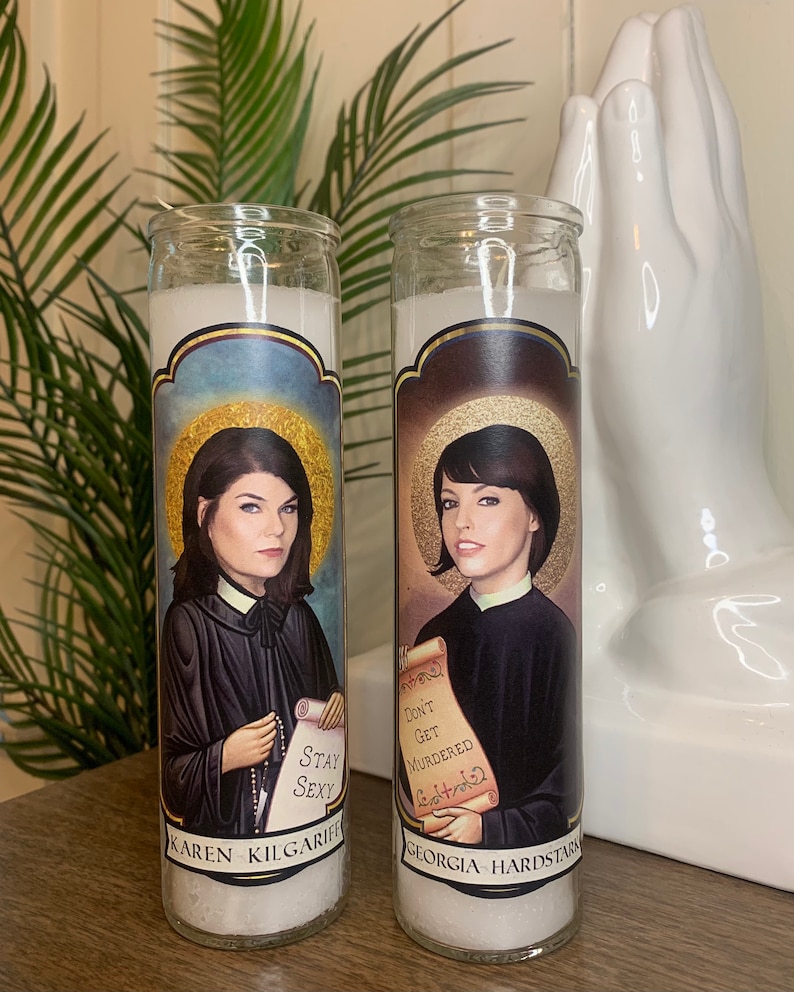 May include: Two white prayer candles with images of women and text. The candle on the left has an image of a woman with dark hair and the text "Karen Kilgariff Stay Sexy". The candle on the right has an image of a woman with dark hair and the text "Georgia Hardstark Don't Get Murdered Eat Pizza".