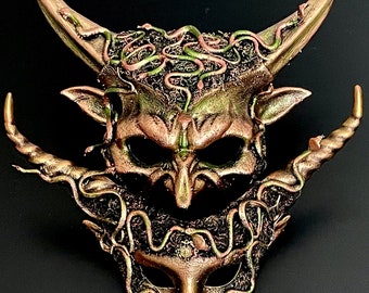 May include: A gold and green masquerade mask with a horned, demonic face and intricate snake designs. The mask is made of a hard material and has a detailed, textured finish.