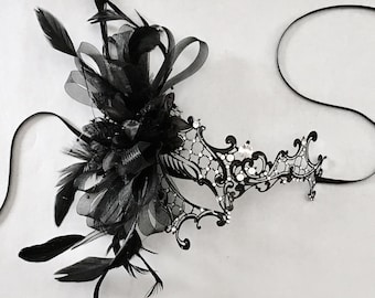 May include: A black masquerade mask with a delicate lace design and rhinestones. The mask is adorned with a black feather and tulle bow.