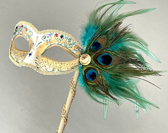 May include: A decorative masquerade mask with a cream-colored base, adorned with colorful rhinestones and gold trim. The mask features a handle and is embellished with peacock feathers in shades of teal and green.