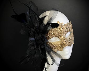 May include: A gold masquerade mask with black lace and a black feather and flower embellishment. The mask has a single eye opening and is decorated with rhinestones.