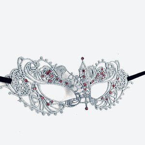 May include: A silver masquerade mask with intricate lace detailing and pink rhinestones. The mask has a black elastic band.