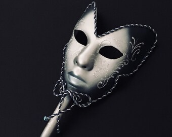 Könnte beinhalten: Eine silberne Maskenballmaske mit einem schwarz-weißen Band, das an einem Stock befestigt ist. Die Maske hat ein filigranes Design mit silbernem Glitzer und einem weißen Gesicht.