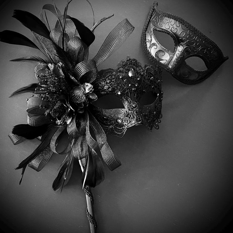 May include: Two black masquerade masks. One mask is embellished with feathers, ribbons, and a decorative handle. The other mask is a simple eye mask with an ornate design. The masks are set against a dark background.