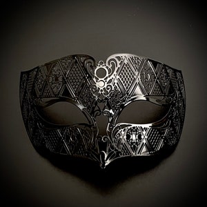 May include: A black, intricately designed masquerade mask. The mask features a symmetrical pattern of diamond shapes and floral filigree, with eye openings and a pointed lower edge. The mask is made of a shiny, reflective material.