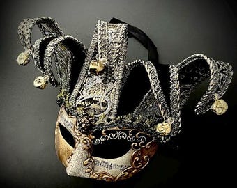 Könnte beinhalten: Eine Maskenballmaske im Narrenstil mit einer mehrzackigen Krone aus silber-schwarz gemustertem Stoff. Das Gesicht der Maske zeigt eine cremefarbene Rissoptik mit Notenaufdrucken, goldenen Filigranverzierungen und kleinen goldenen Glöckchen.