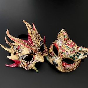 May include: Two ornate Venetian masquerade masks, one with a dragon design and the other with a cat design. Both masks are painted in gold, red, and green with intricate details.