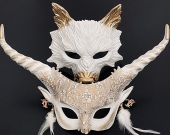 May include: Two white masquerade masks with gold accents. The top mask is a wolf with pointed ears and a textured face. The bottom mask is a horned creature with intricate details and feathers.