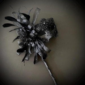 May include: A black masquerade mask with intricate lace detailing and embellishments. The mask is adorned with black feathers, ribbons, and a decorative handle. The overall design is elegant and suitable for a formal event or costume party.
