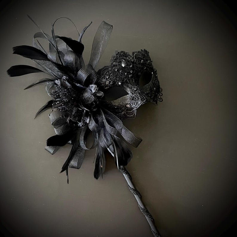 May include: A black masquerade mask with intricate lace detailing and embellishments. The mask is adorned with black feathers, ribbons, and a decorative handle. The overall design is elegant and suitable for a formal event or costume party.