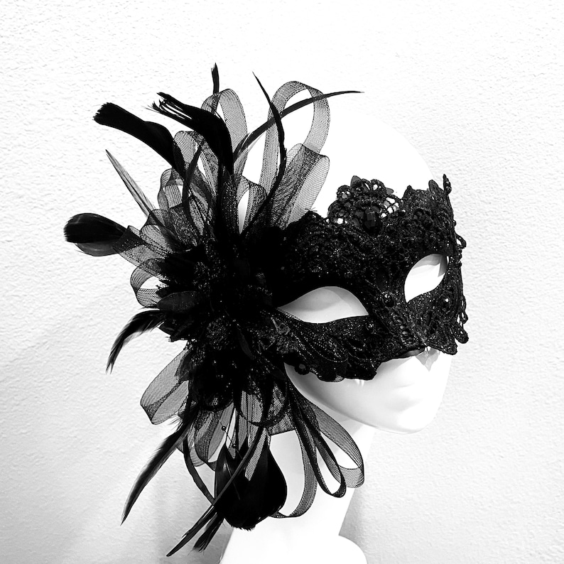 May include: Black lace masquerade mask with feather and ribbon accents. The mask features intricate lace detailing around the eye openings and a decorative arrangement of black feathers and sheer ribbons on the side. The mask is displayed on a white mannequin head.