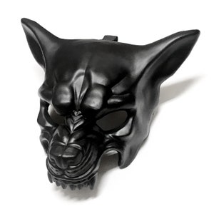 Puede incluir: Una m&aacute;scara de pl&aacute;stico negro moldeado con forma de lobo o perro gru&ntilde;endo. La m&aacute;scara tiene orejas puntiagudas, un hocico detallado y aberturas para los ojos. Se adjunta una correa negra para usarla.