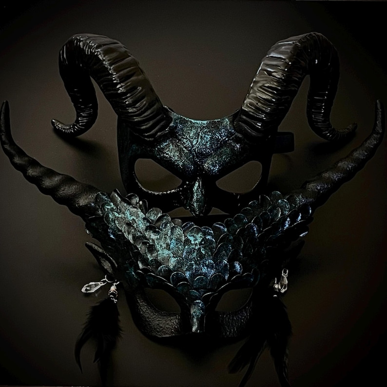 May include: Two black fantasy masks with intricate details. One mask features large, curved horns, while the other has a feathered design and dangling accents. Both masks have a dark, textured finish with hints of teal.