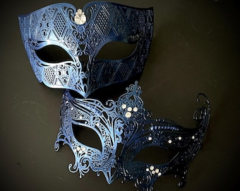 May include: Two dark blue metal masquerade masks featuring intricate laser-cut filigree patterns. The top mask has a geometric design with a single clear crystal, while the bottom mask displays ornate scrollwork accented with multiple clear crystals.