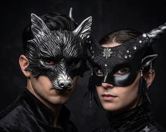 May include: A silver wolf mask with detailed fur texture and a black mask featuring lace, rhinestones, a central emblem, and black feathers. Both masks are worn over the eyes, with the wolf mask covering the upper face and the lace mask featuring horns.