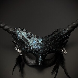 May include: A black fantasy mask with large, curved horns and a feathered design. The mask features iridescent scales and dangling silver accents. Black feathers extend from the bottom of the mask, creating a dramatic effect.