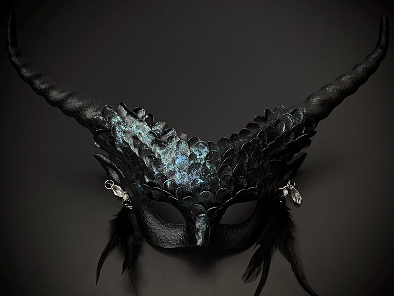May include: A black fantasy mask with large, curved horns and a feathered design. The mask features iridescent scales and dangling silver accents. Black feathers extend from the bottom of the mask, creating a dramatic effect.