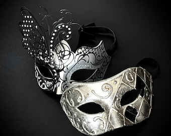 May include: Two decorative masquerade masks. One mask is silver with black accents and a butterfly wing design. The other mask is silver with a harlequin pattern. Both masks have black ribbons for securing them.