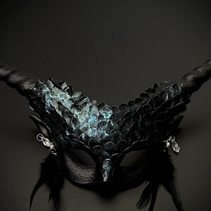 May include: A black fantasy mask with large, curved horns and intricate detailing. The mask features a textured surface with iridescent blue-green accents, small feathers, and dangling crystal charms, creating a dramatic and mysterious look.
