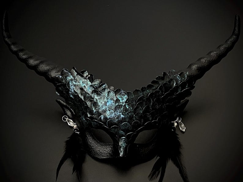 May include: A black fantasy mask with large, curved horns and intricate detailing. The mask features a textured surface with iridescent blue-green accents, small feathers, and dangling crystal charms, creating a dramatic and mysterious look.