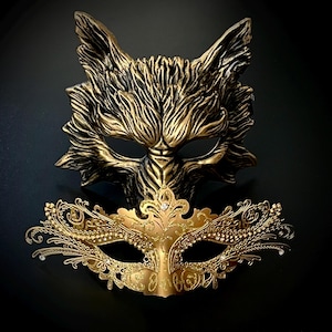 May include: Two gold masquerade masks. One mask is a detailed wolf design, the other is a delicate, ornate design with rhinestones.