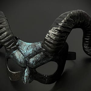 May include: A black horned mask with a textured surface and a hint of blue-green highlights. The mask features large, curved horns and eye openings, connected by a black elastic band. The mask is designed for costume or decorative purposes.