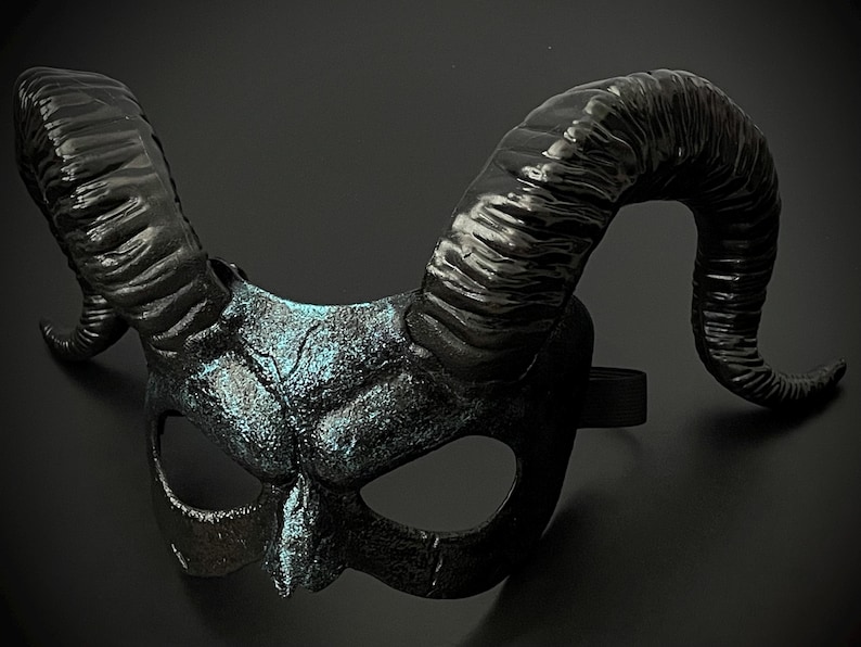 May include: A black horned mask with a textured surface and a hint of blue-green highlights. The mask features large, curved horns and eye openings, connected by a black elastic band. The mask is designed for costume or decorative purposes.