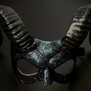 May include: A black horned mask with a textured finish and iridescent blue highlights. The mask features large, curved horns and eye openings, suitable for costume or theatrical use.