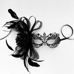 May include: A black masquerade mask with intricate lace detailing and rhinestones. The mask has a black feather and tulle embellishment attached to the side.