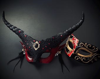 May include: Two ornate masquerade masks. One is a devil-horned mask in black and red with jewel accents and feathers. The other is a smaller mask in black, red, and gold with a decorative swirl design.