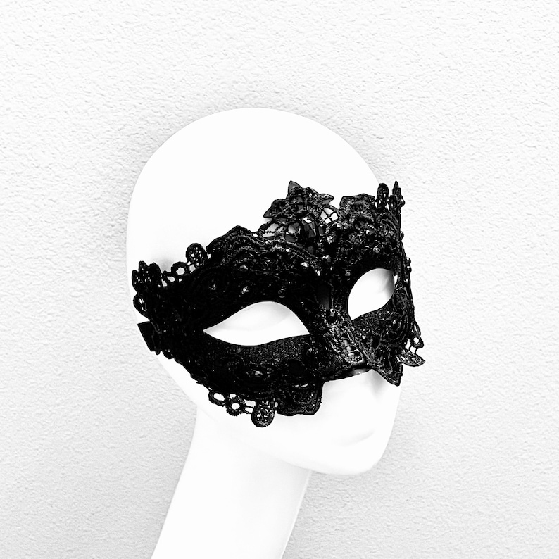 May include: A black lace masquerade mask with intricate floral and scrollwork designs. The mask covers the eyes and features a delicate, scalloped edge. The mask is displayed on a white mannequin head.