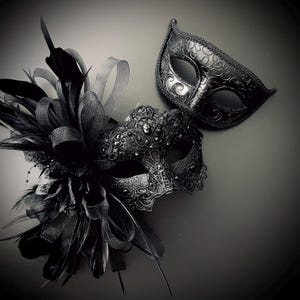 May include: Two black masquerade masks. One mask is embellished with feathers, ribbons, and beads. The other mask has a simple, elegant design. Both masks are made of a dark material and are suitable for a formal event or costume party.