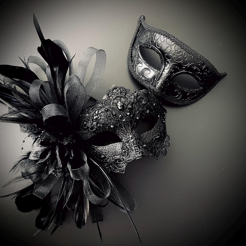 May include: Two black masquerade masks. One mask is embellished with feathers, ribbons, and beads. The other mask has a simple, elegant design. Both masks are made of a dark material and are suitable for a formal event or costume party.