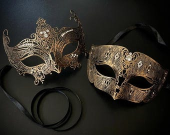 May include: Two ornate masquerade masks in a metallic bronze finish with intricate filigree and geometric cutouts. Both masks feature clear crystal accents and are attached to black satin ribbons for securing around the head.